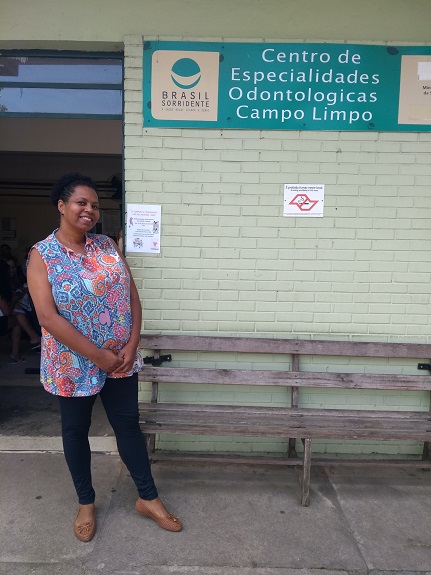 Ana Maria posa em frente ao Centro de especialidades Odontológicas Campo Limpo. Ao centro, há um banco de madeira.